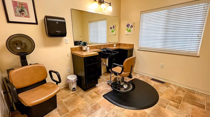 Small salon-style room with two brown styling chairs, a hair-washing sink, mirror and countertop beneath a window.