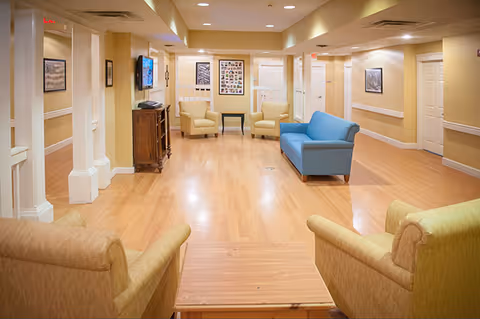 Bright common lounge with sofas and armchairs arranged on a wood floor in a senior living facility.