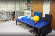 Physical therapy room with a blue padded treatment table holding exercise equipment including a yellow exercise ball, small dumbbells, and foam rollers. In the background, there are walkers lined up against the wall and a wooden door with mounted exercise bars.