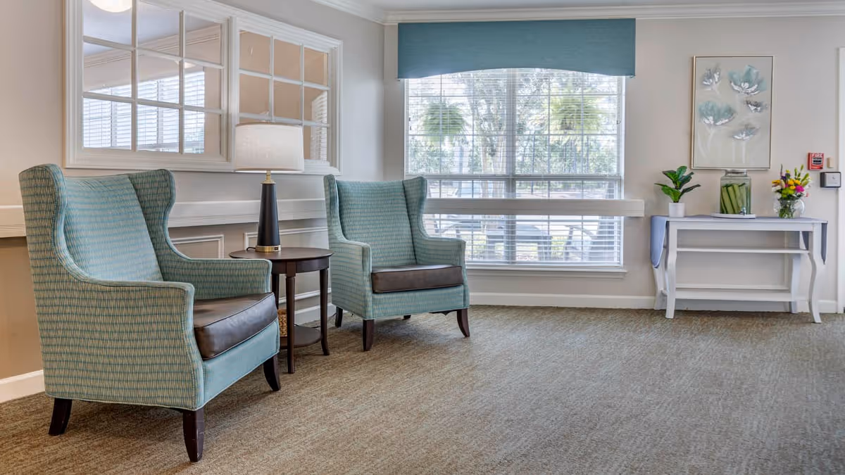 Two upholstered armchairs and a small table with a lamp in a bright seating area by a large window and a console table with plants and artwork.