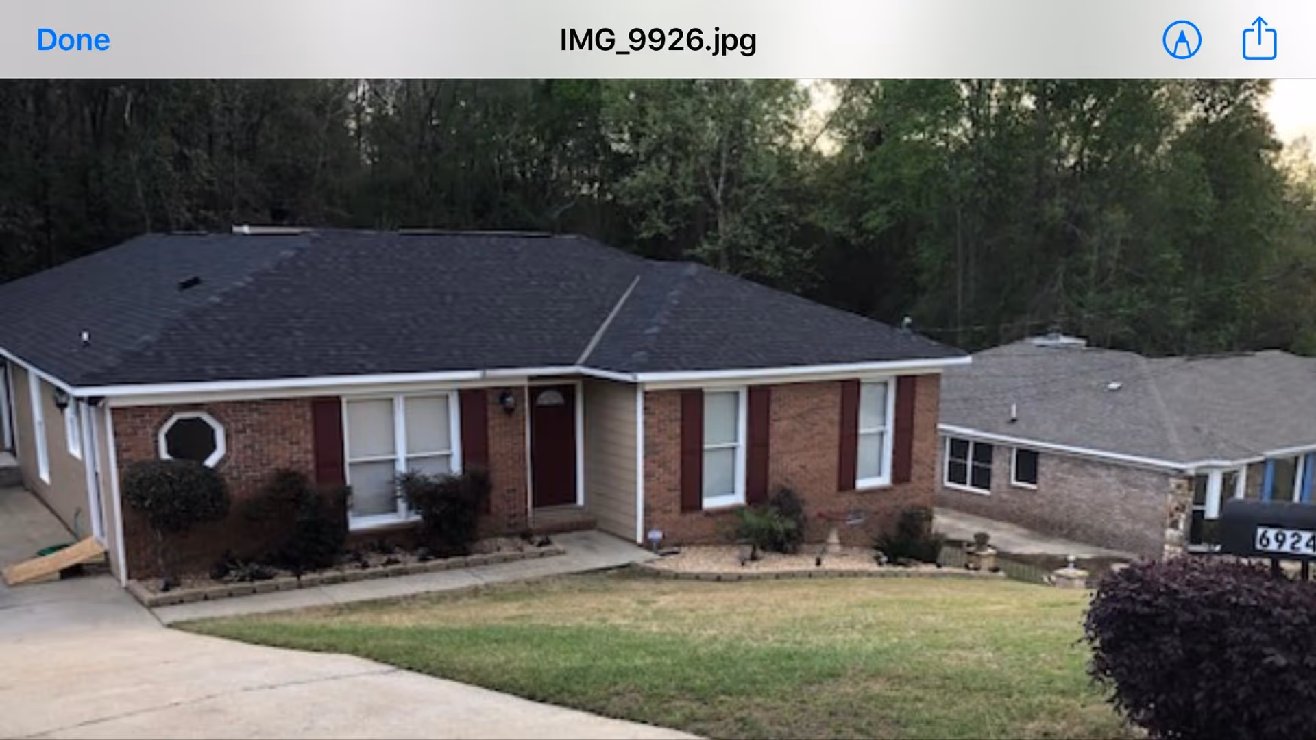 Single-story brick house with a dark shingled roof, red front door, and several windows with red shutters. The house is surrounded by a small garden with shrubs and a sloped lawn. Another similar house is visible in the background, along with trees.