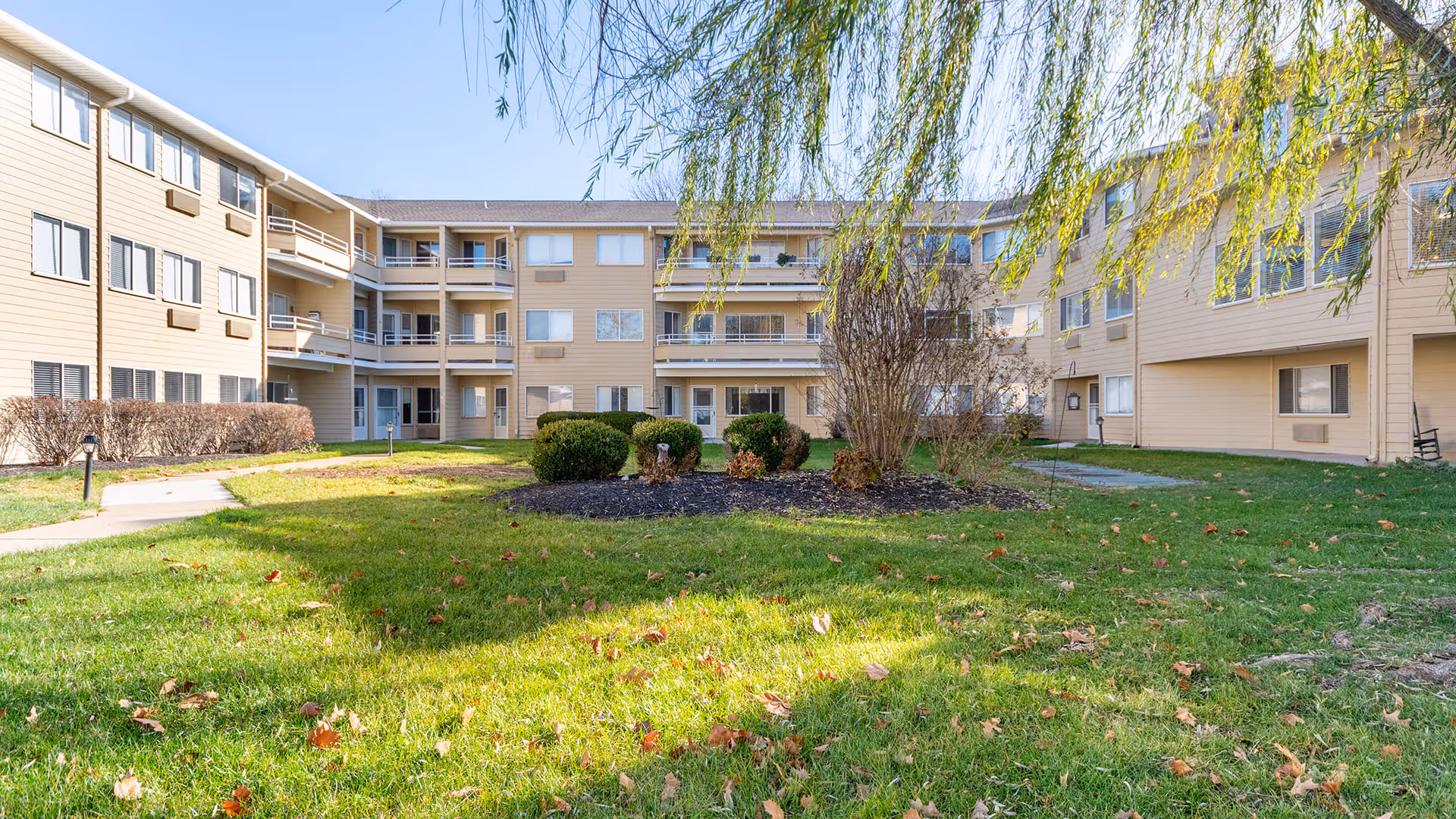 Outdoor courtyard area of a senior living facility with green grass, bushes, and a few leafless trees surrounded by a three-story beige building with balconies and windows under a clear blue sky.