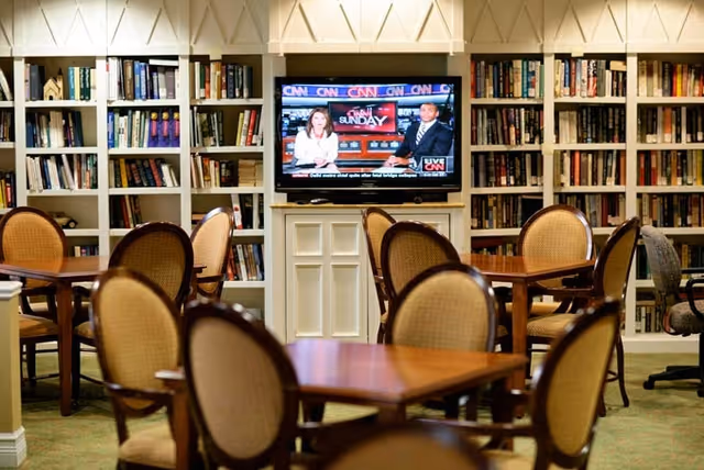 Community lounge with tables and chairs facing a wall of bookshelves and a television displaying a news broadcast.