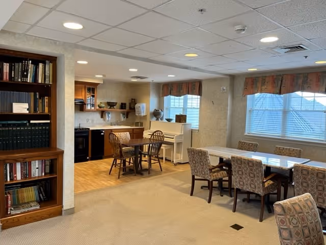 Communal dining and activity room with multiple tables and chairs, a small kitchenette, bookshelf, and an upright piano.
