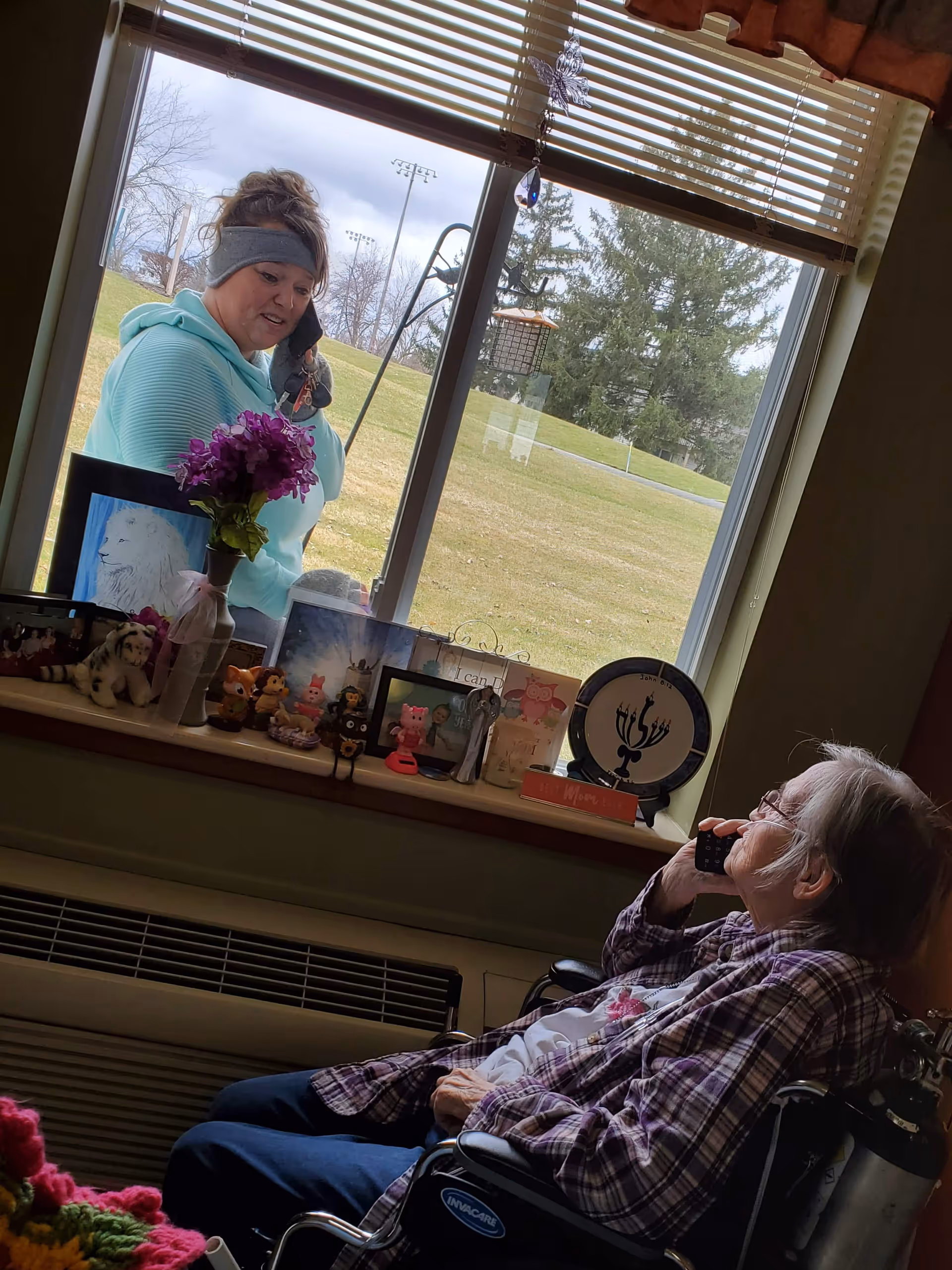 An elderly woman in a wheelchair inside a room talking on a phone, looking out a window where a younger woman is also talking on a phone outside. The window sill is decorated with various small figurines, framed pictures, a vase with purple flowers, and a decorative plate. Outside, there is a grassy area with trees and a bird feeder visible.