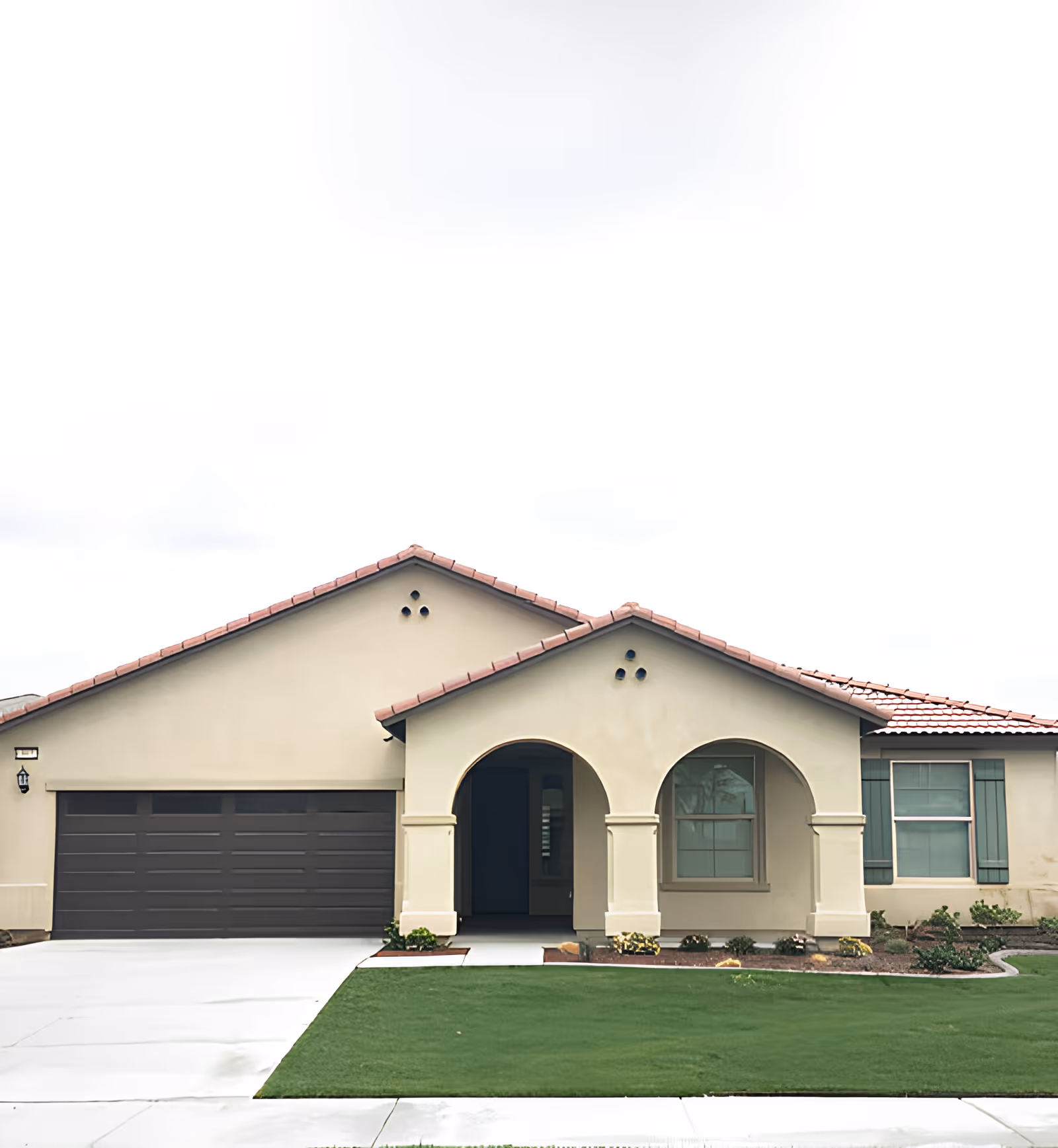 Front exterior view of a single-story house with a beige stucco finish, red tile roof, two arched entryways, a dark brown garage door, and green lawn with some landscaping.