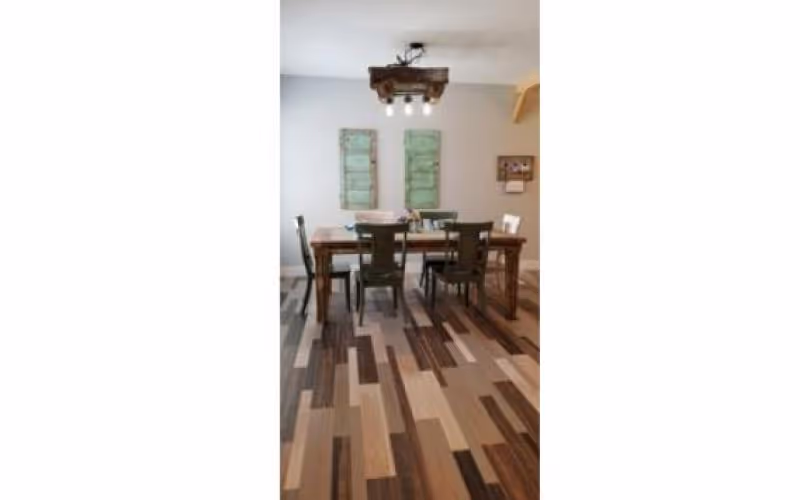 A dining area with a wooden table and four chairs on a patterned wooden floor. Two green decorative panels hang on the white wall behind the table, and a rustic light fixture with exposed bulbs is mounted on the ceiling above the table.