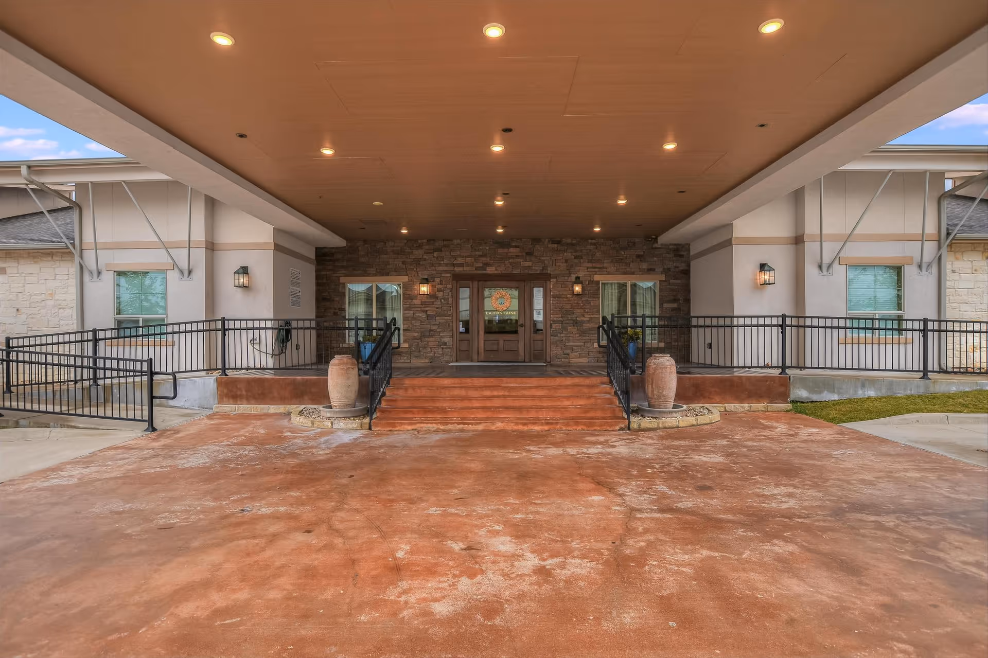 Covered main entrance of a memory care facility with a wide driveway, steps and ramps leading to double doors set in a stone facade.