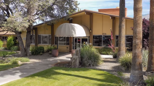 Exterior view of Pueblo Springs Rehabilitation Center showing a single-story building with a beige facade, large windows, an entrance with a white canopy, surrounded by green grass, trees, and landscaping including bushes and palm trees.