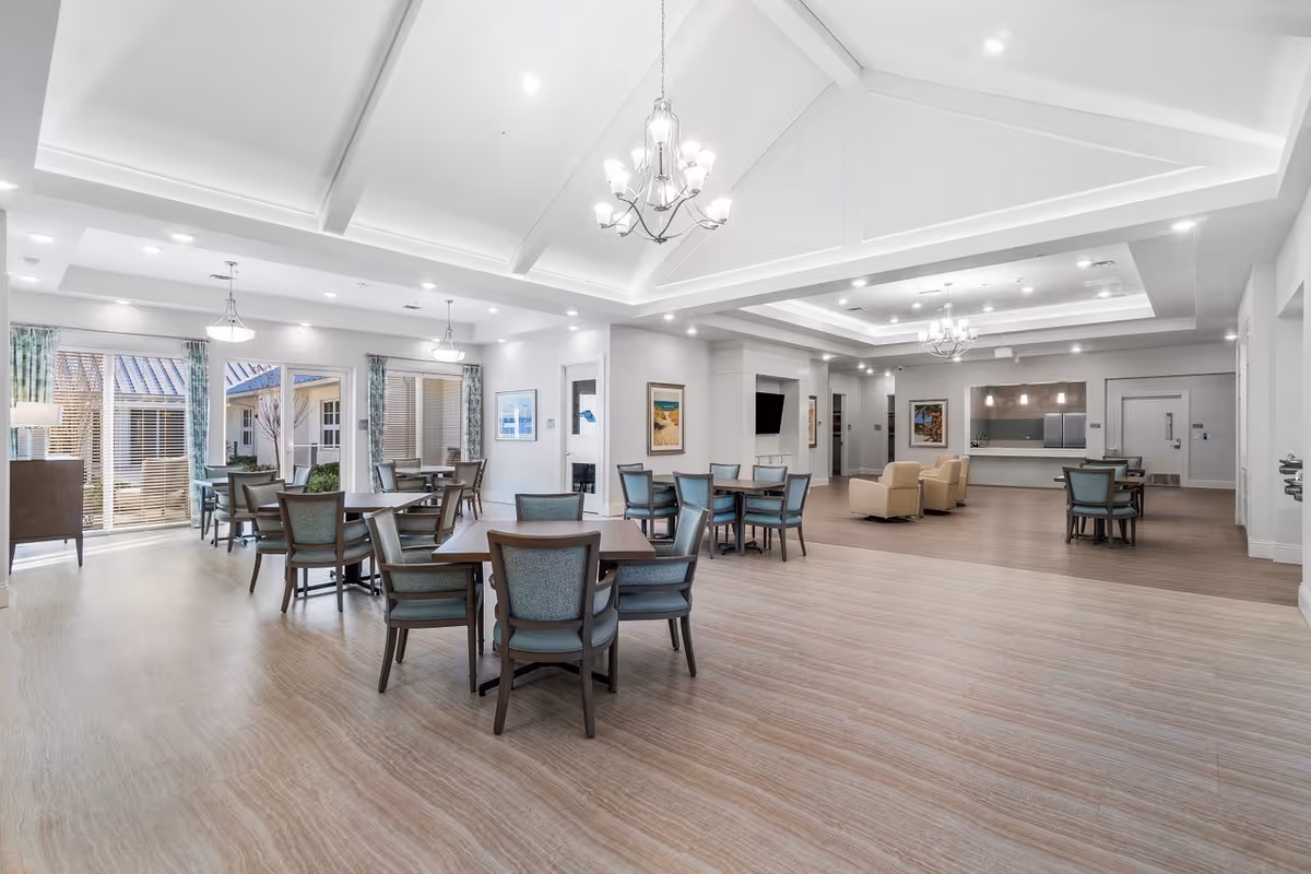 Bright, open communal dining and lounge area with multiple tables and chairs, chandeliers, high ceilings, and large windows.