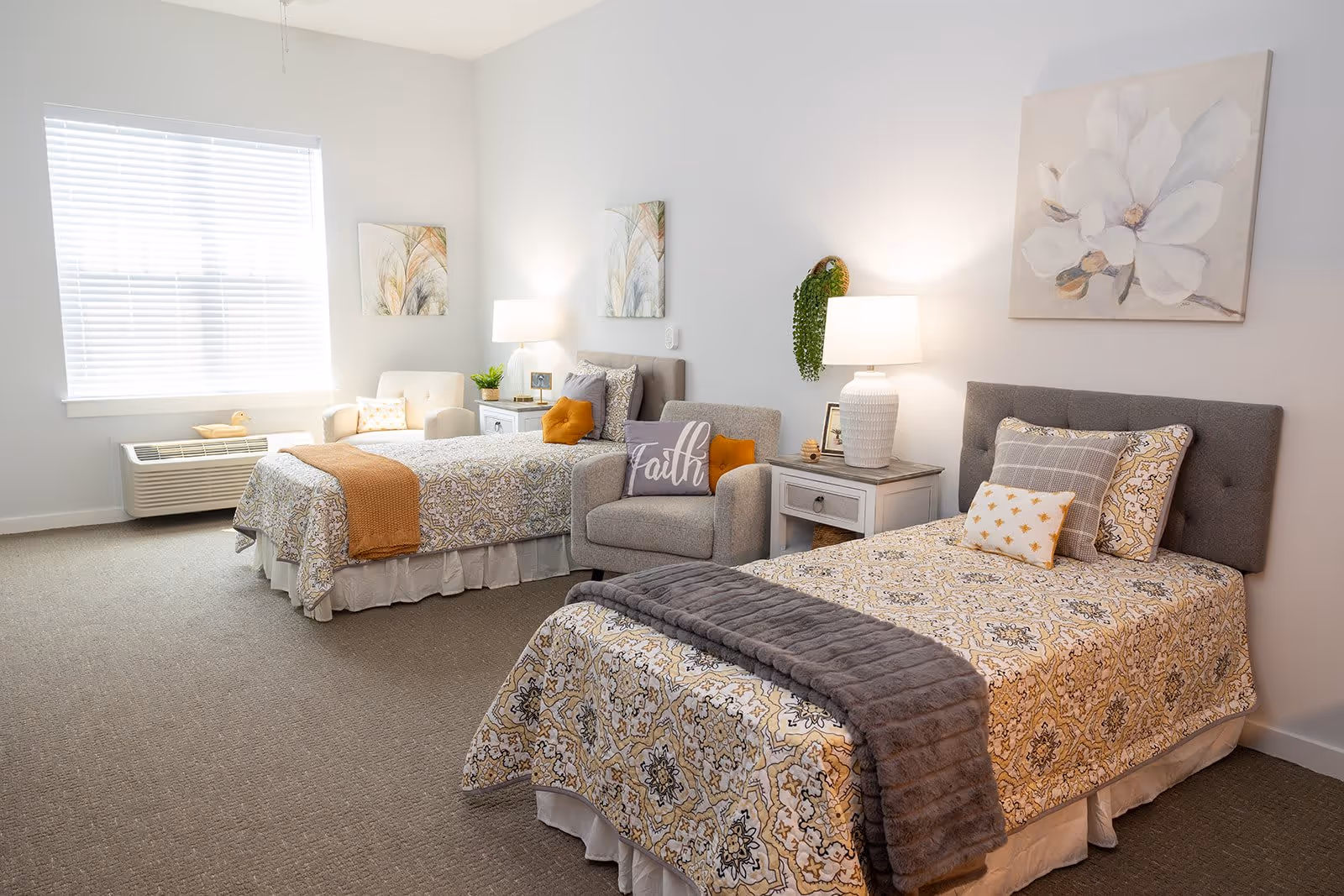 A bright and cozy bedroom with two twin beds, each adorned with patterned bedspreads and decorative pillows. Between the beds is a nightstand with a white lamp and small decor items. A comfortable armchair with a 'Faith' pillow is placed near one bed. The room has light-colored walls, a large window with blinds, and framed artwork on the walls.