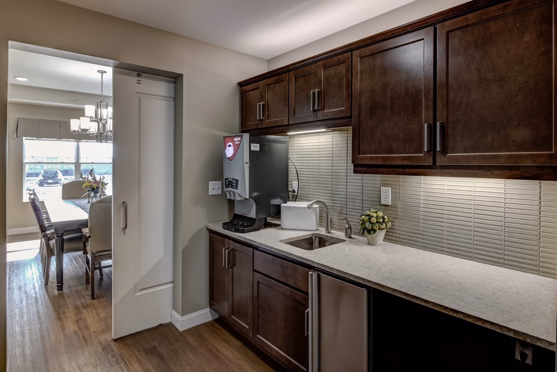 Small kitchenette with dark wood cabinets, a countertop sink and undercabinet lighting, opening to a dining area through a doorway.