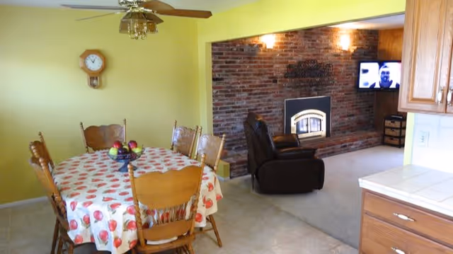 Interior view of a senior living facility showing a dining area with a round table covered with a fruit-patterned tablecloth and six wooden chairs. A ceiling fan with lights is above the table. Adjacent to the dining area is a living room with a brick fireplace, a black recliner chair, and a wall-mounted TV. The walls are painted yellow, and part of a kitchen counter with wooden cabinets is visible on the right.