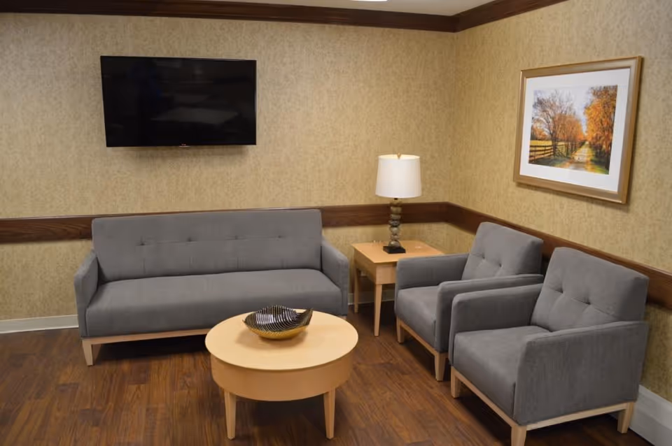 A cozy living room area with a gray sofa, two matching gray armchairs, a round wooden coffee table with a decorative bowl, a wooden side table with a lamp, a wall-mounted flat-screen TV, and a framed picture of a tree-lined path on the wall.