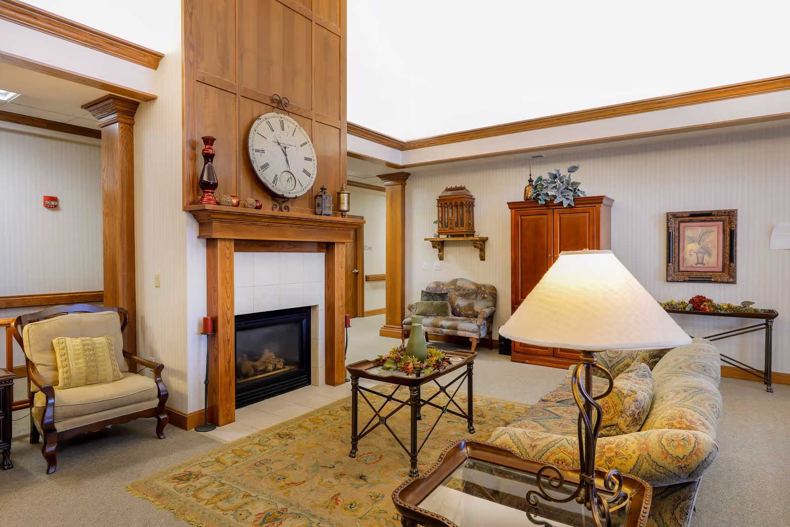 A cozy senior living facility common area with a large wooden fireplace featuring a clock above it. The room includes a patterned sofa, an armchair with a yellow cushion, a glass-top coffee table with decorative items, a wooden cabinet, and a framed picture on the wall. The space is warmly lit with a table lamp and has beige carpeting and light-colored walls with wood trim.