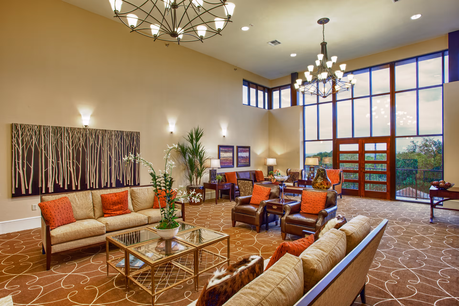 A spacious and well-lit living room with high ceilings and large windows. The room features multiple seating areas including beige sofas with orange cushions, brown leather armchairs, and wooden side tables. There are decorative plants, modern wall art, and elegant chandeliers hanging from the ceiling. The carpet has a circular pattern, and the large windows offer a view of greenery outside.