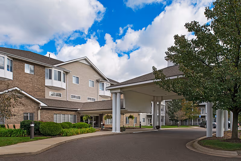 Covered entrance and porte-cochere of a multi-story senior living building with landscaping and a curved driveway under a partly cloudy sky.