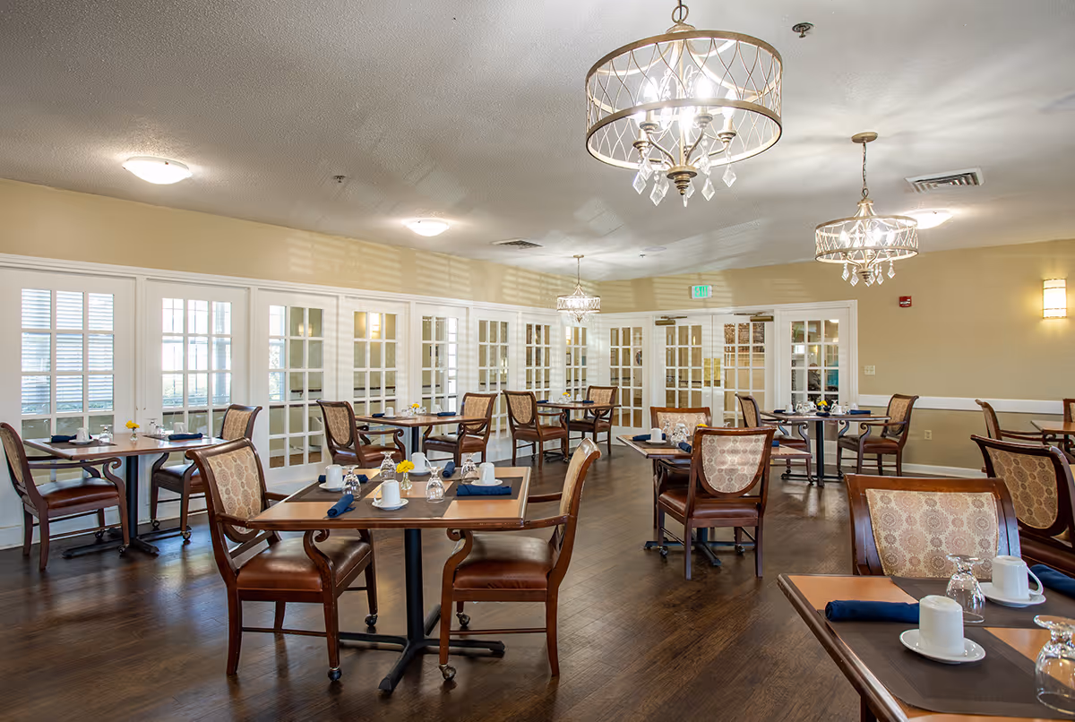 A well-lit dining room with multiple wooden tables and chairs arranged neatly. Each table is set with white cups, glasses, and blue napkins. The room features large windows with white frames and glass-paneled doors. Elegant chandeliers hang from the ceiling, and the floor is made of dark wood.
