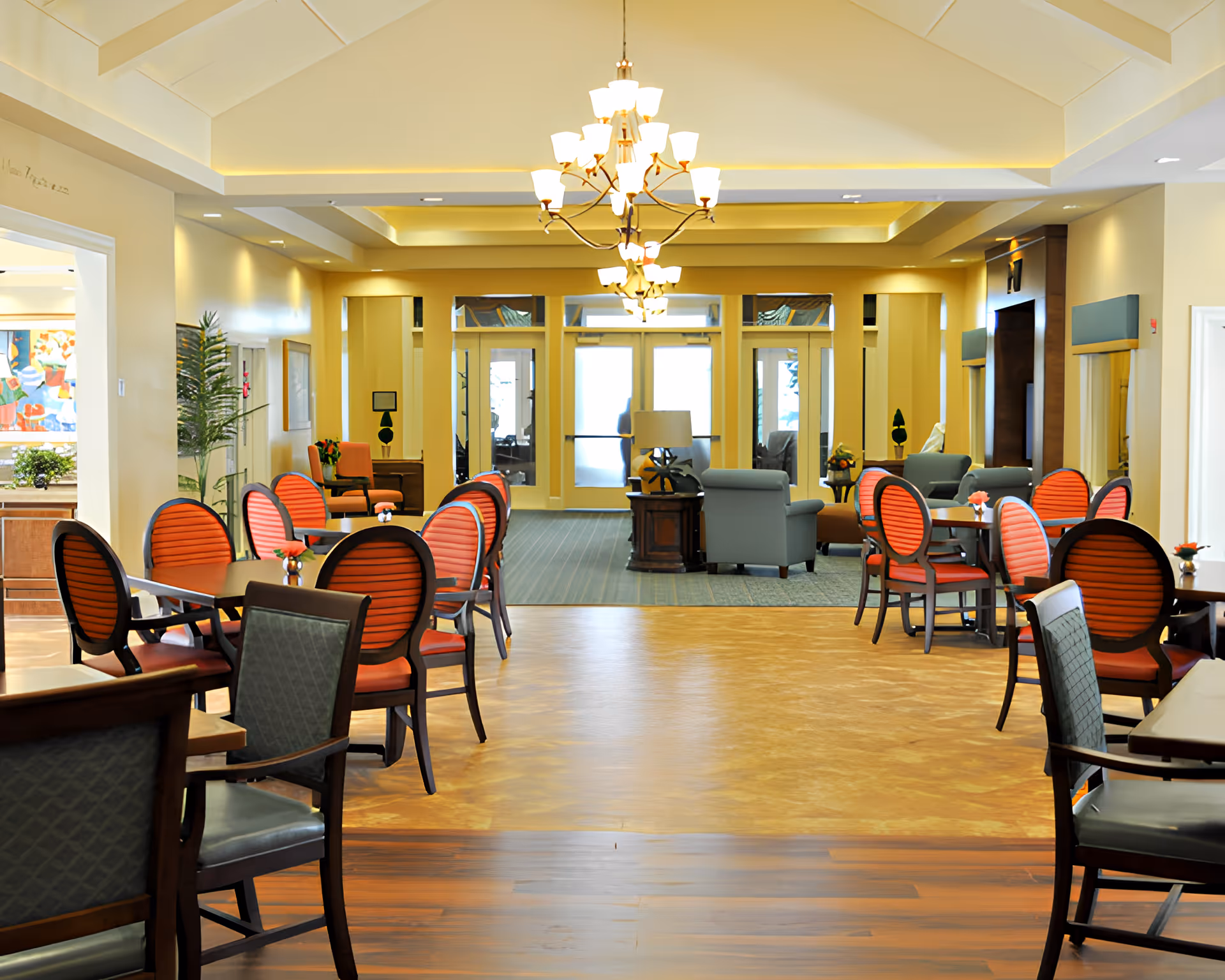 A spacious and well-lit common area in a senior living facility featuring multiple tables with red cushioned chairs arranged neatly. The room has a high ceiling with elegant chandeliers, and in the background, there is a seating area with armchairs and a lamp near large glass doors.