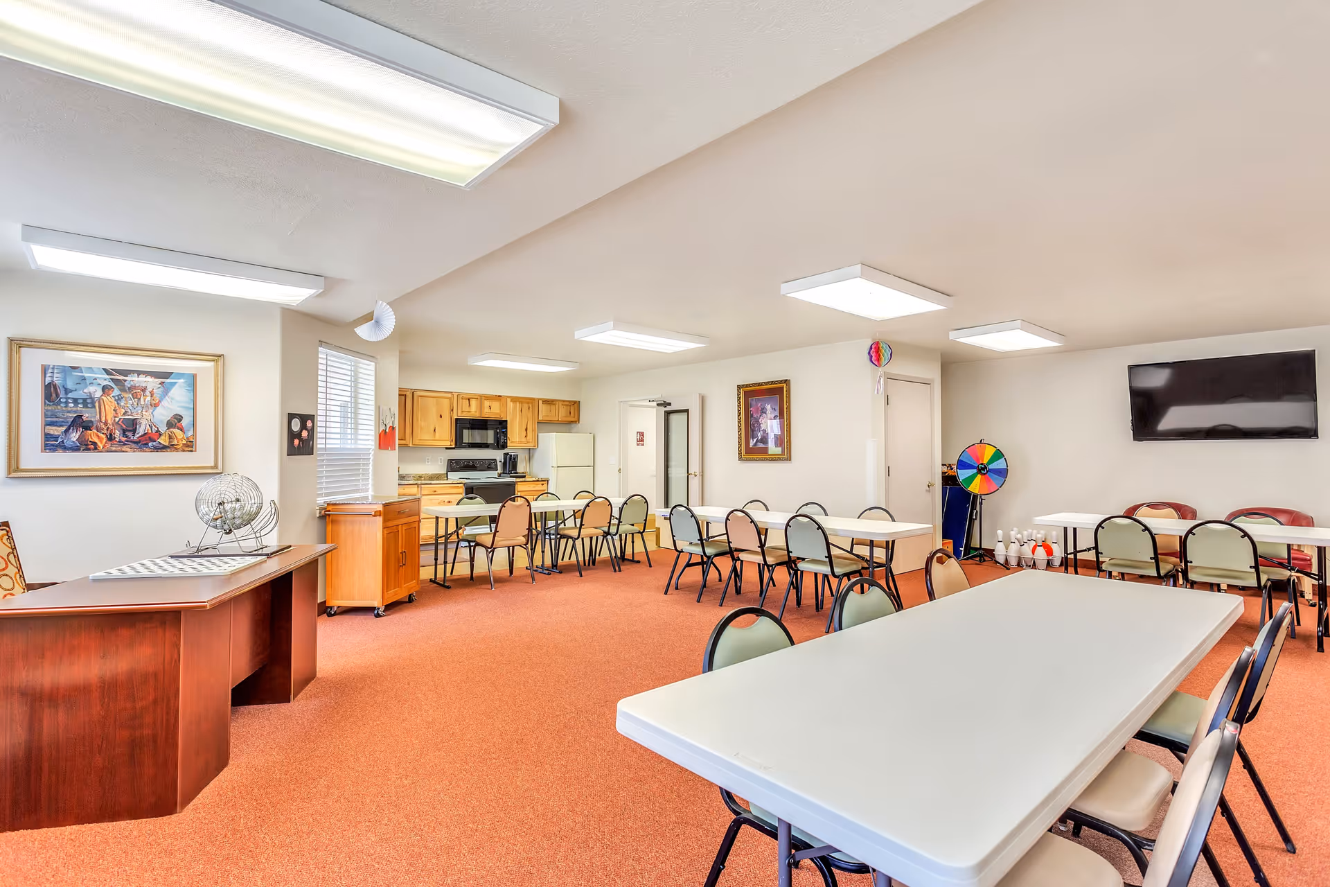 A spacious common room with multiple tables and chairs arranged for group activities or dining. The room features a kitchenette area with wooden cabinets, a microwave, stove, and refrigerator. There is a large flat-screen TV mounted on the wall, colorful game wheels, and framed artwork on the walls. The floor is carpeted in a reddish-orange color, and the ceiling has several rectangular fluorescent light fixtures.