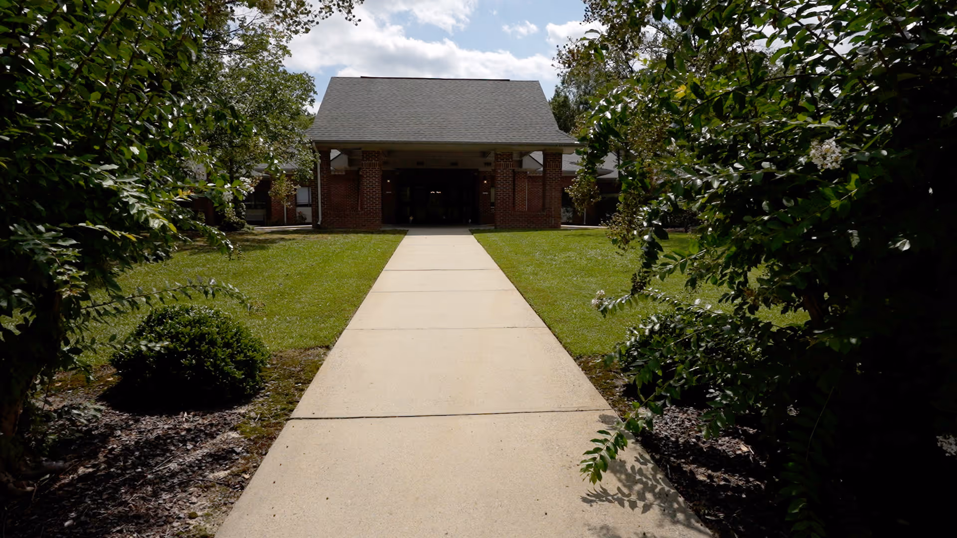 A concrete walkway leading through a well-maintained grassy area with bushes and trees on either side, heading towards the entrance of a brick building with a covered porch and a pitched roof under a partly cloudy sky.