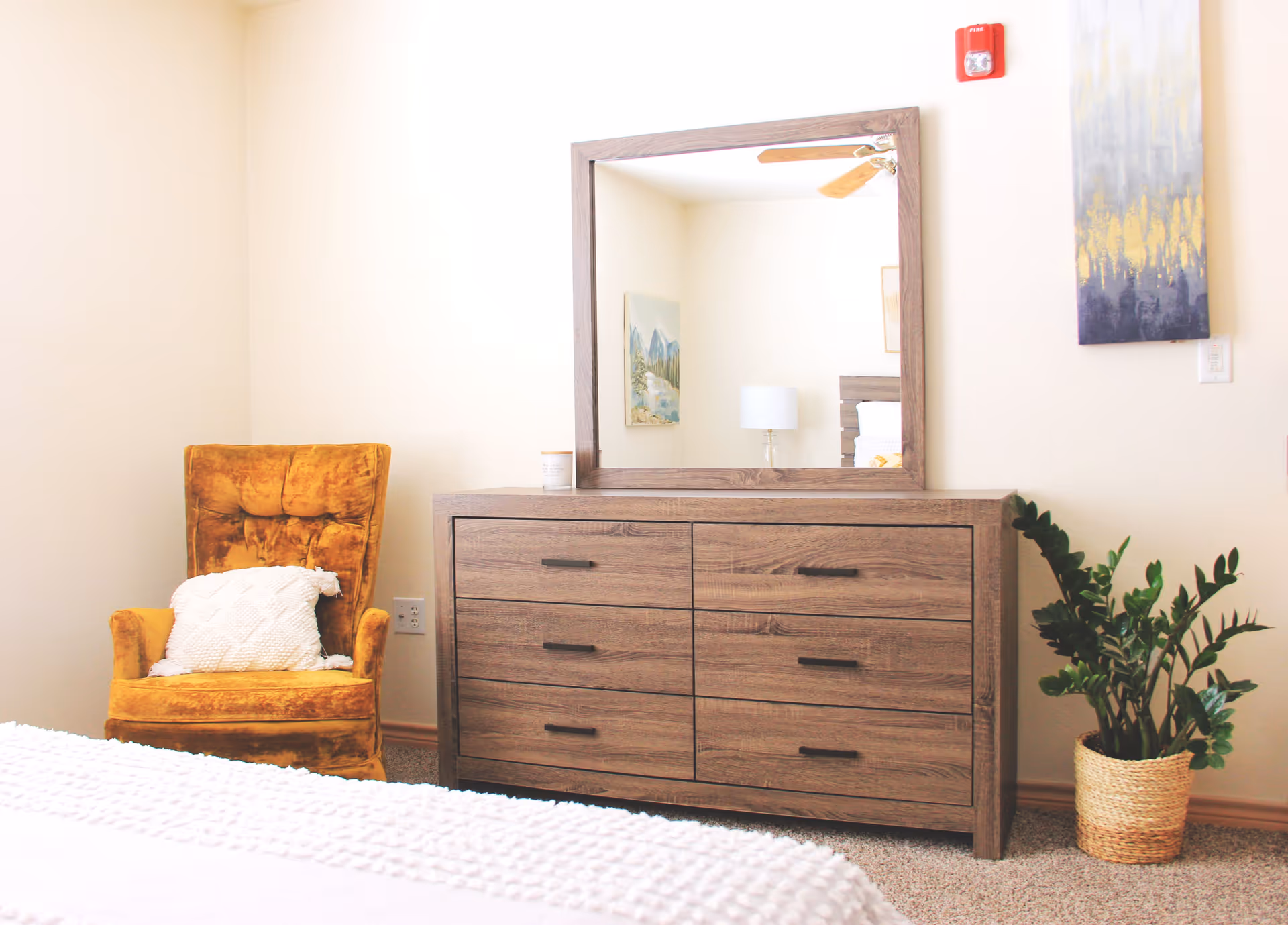A cozy bedroom corner featuring a mustard yellow upholstered armchair with a white textured pillow, a wooden dresser with six drawers and a large rectangular mirror on top, a potted green plant in a woven basket, and light-colored walls adorned with two pieces of artwork.