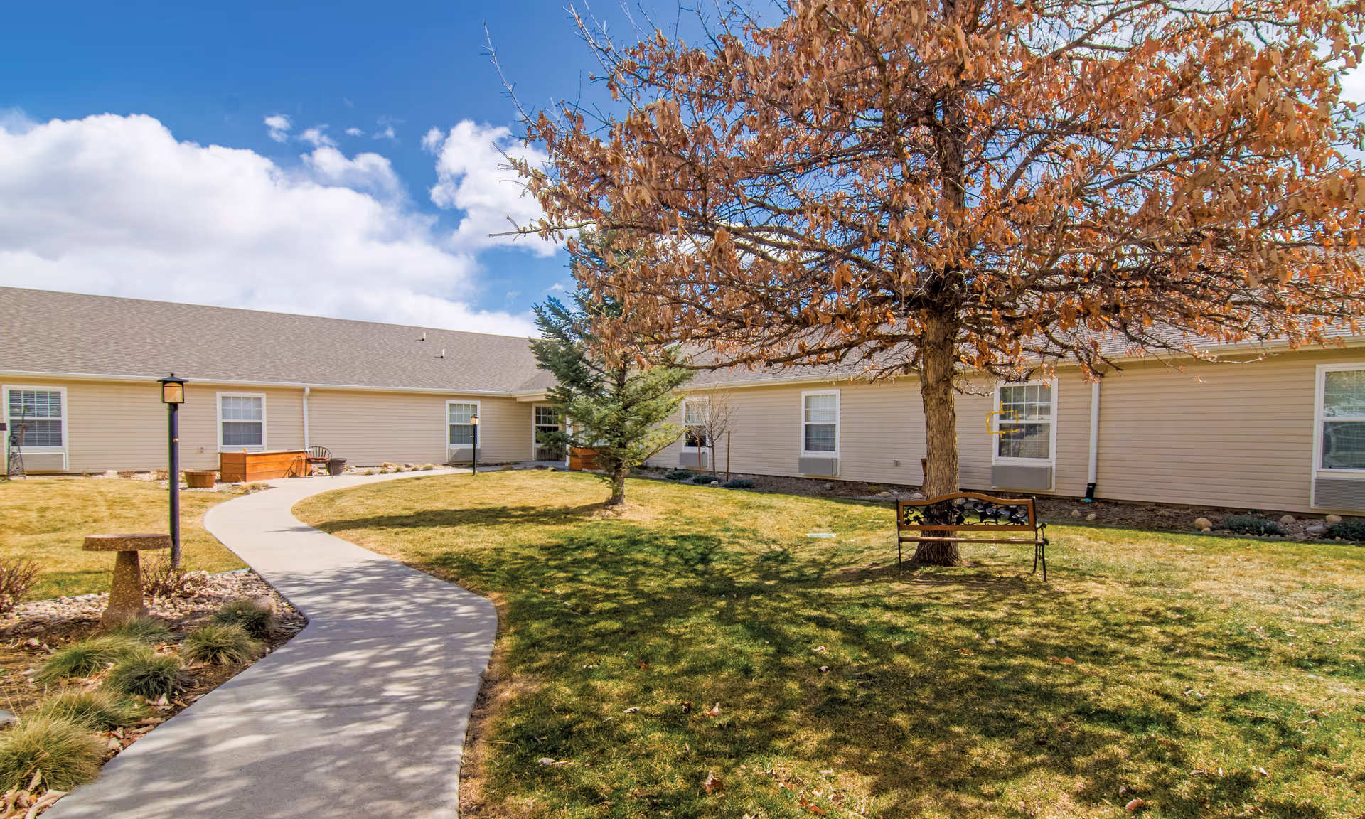 Outdoor courtyard area with a curved concrete walkway, a bench under a tree with brown leaves, green grass, and a beige building with multiple windows in the background under a partly cloudy blue sky.