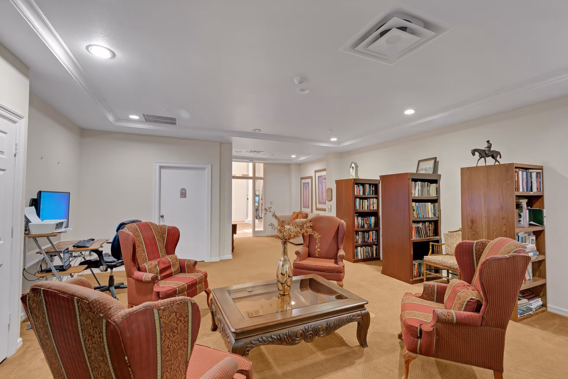 A cozy common area in Asher Point Independent Living of Sherman featuring four upholstered armchairs arranged around a wooden coffee table with a decorative vase. The room has beige walls and carpet, bookshelves filled with books, framed artwork on the walls, and a computer workstation in the corner.