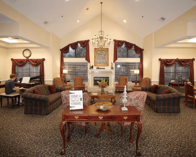 A spacious and warmly decorated living room in a senior living facility with high vaulted ceilings and a chandelier. The room features multiple upholstered armchairs and sofas arranged around a central coffee table with a flower vase. A fireplace with a 'Welcome' sign above it is centered between two large windows with red curtains. A man is playing a piano on the left side of the room. A wooden table with decorative items and a sign that reads 'Home Sweet Home' is in the foreground.