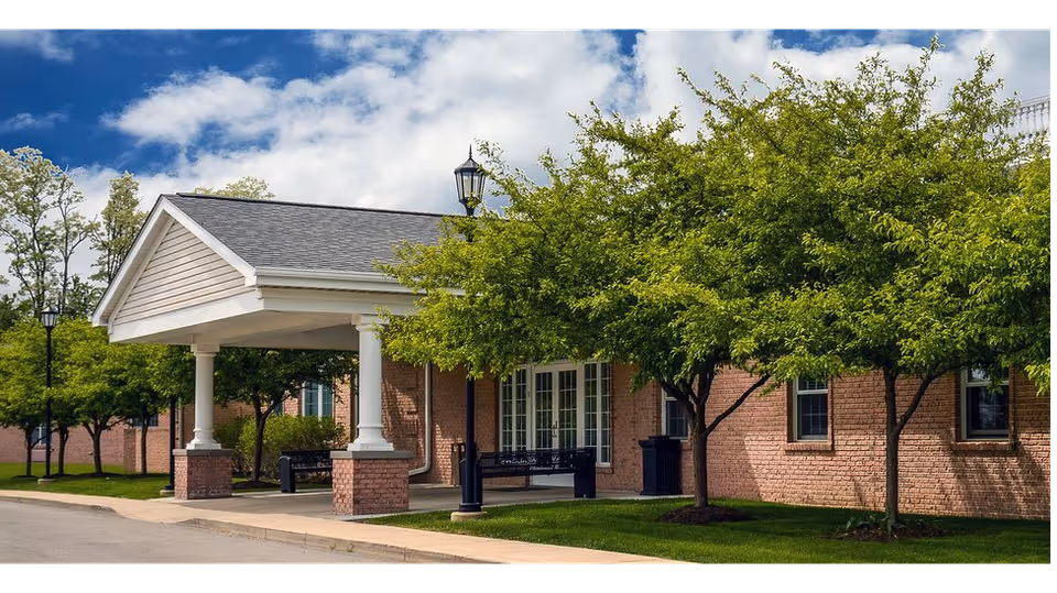 Exterior view of a brick building with a covered entrance supported by white columns. There are green trees and a bench near the entrance, with a clear blue sky and some clouds above.