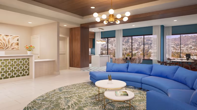 Bright memory care common lounge with a curved blue sectional sofa, round coffee tables, a reception desk, and large windows showing a hillside view.