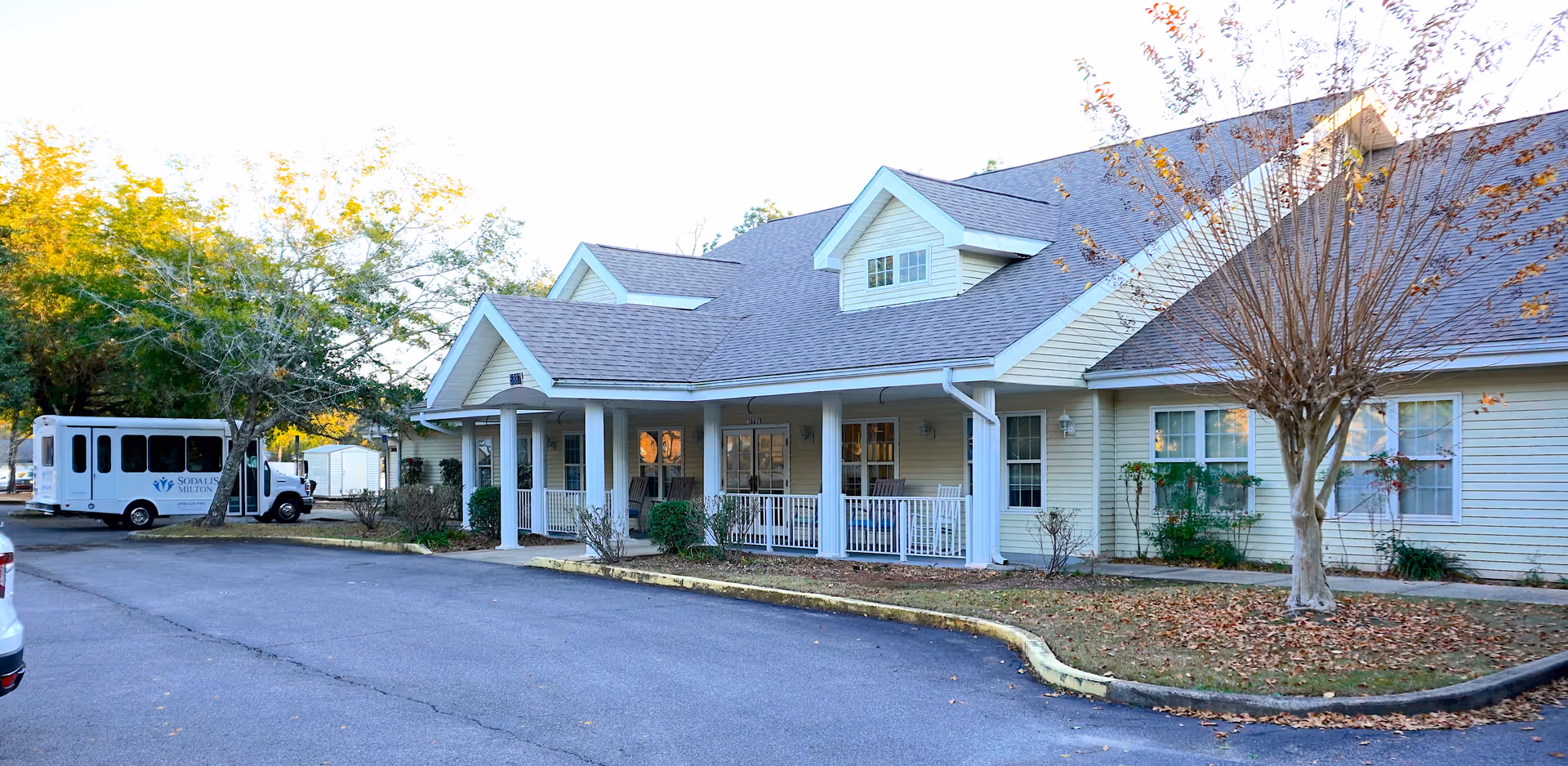Exterior view of a single-story senior living facility building with a covered porch, white railings, and several windows. There is a small parking area in front with a white shuttle bus parked on the left side. Trees with autumn leaves surround the building.