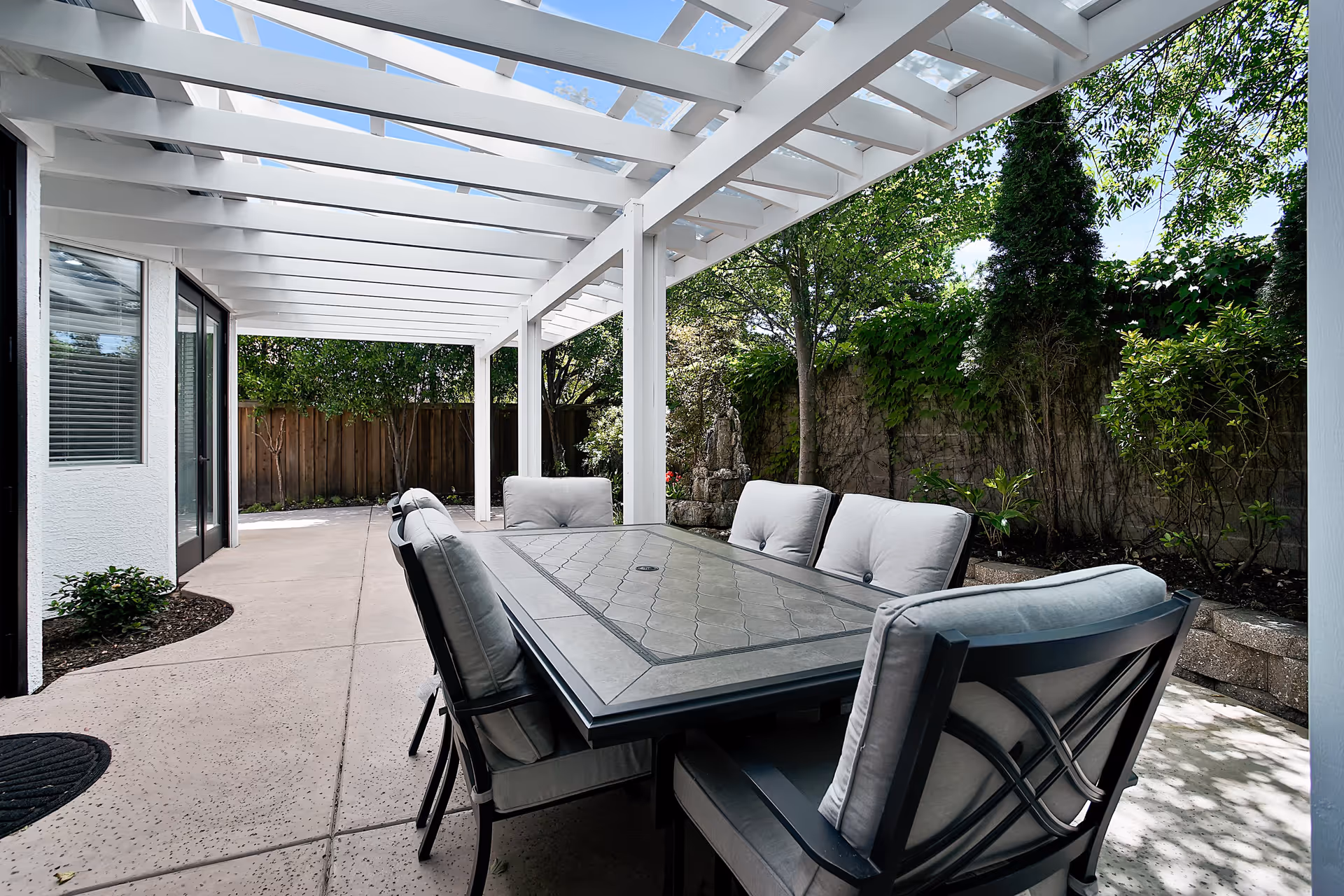 Outdoor patio area with a large rectangular table surrounded by cushioned chairs under a white pergola. The patio is adjacent to a building with glass doors and windows, and is bordered by a wooden fence and greenery including trees and shrubs.