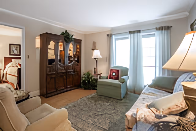 Cozy living room with armchairs, a sofa, a wooden cabinet, a floor lamp, and a large window with sheer curtains.
