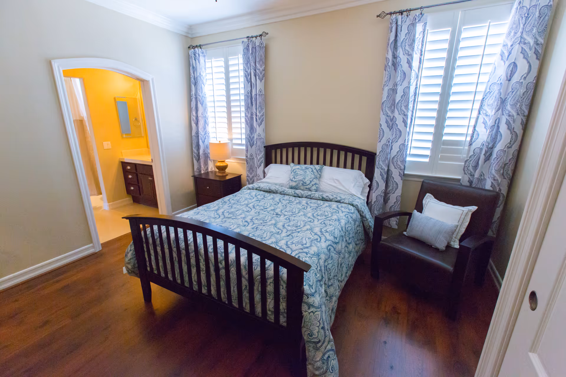 A bedroom with a wooden bed frame and a bed made with blue and white patterned bedding. There is a nightstand with a lamp next to the bed, two windows with white shutters and blue patterned curtains, and a dark brown armchair with two pillows. An open doorway leads to a bathroom with a yellow wall and a vanity with a mirror.