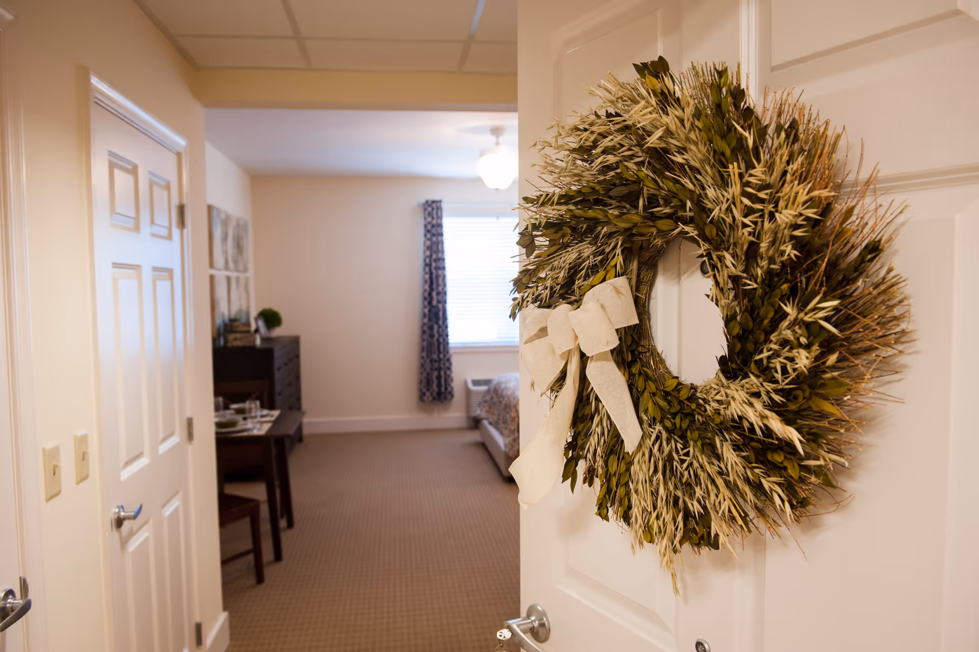 A decorative wreath hangs on a partially open door leading into a furnished senior living apartment with a bed, dining table, and window.