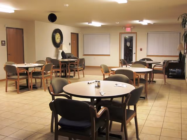 A well-lit dining area with multiple round tables and chairs arranged neatly. The room has tiled flooring, beige walls, and two windows with closed blinds. There is a large clock on one wall, a lamp on a small table, and an exit door with a red exit sign above it.