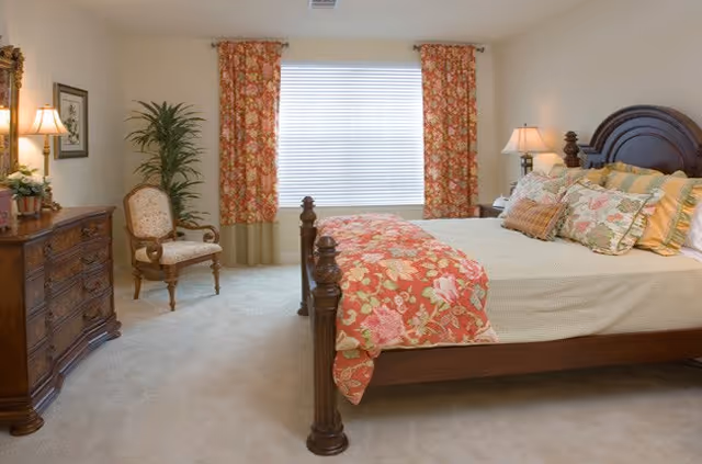 A cozy bedroom with a large wooden bed featuring floral bedding and multiple pillows. There are matching floral curtains on the window behind the bed. To the left, there is a wooden dresser with a lamp and framed artwork above it, and an upholstered armchair next to a tall green plant. The room is softly lit with a bedside lamp on the right side of the bed.