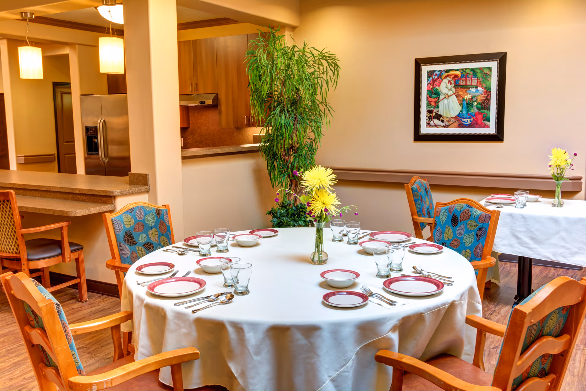 A dining area with a round table covered with a white tablecloth set for six people with plates, bowls, glasses, and silverware. There are wooden chairs with colorful leaf-patterned cushions around the table. In the background, there is a kitchen area with a refrigerator and cabinets, a tall green plant, and a framed colorful painting on the wall. Another smaller table with similar settings is visible to the right.