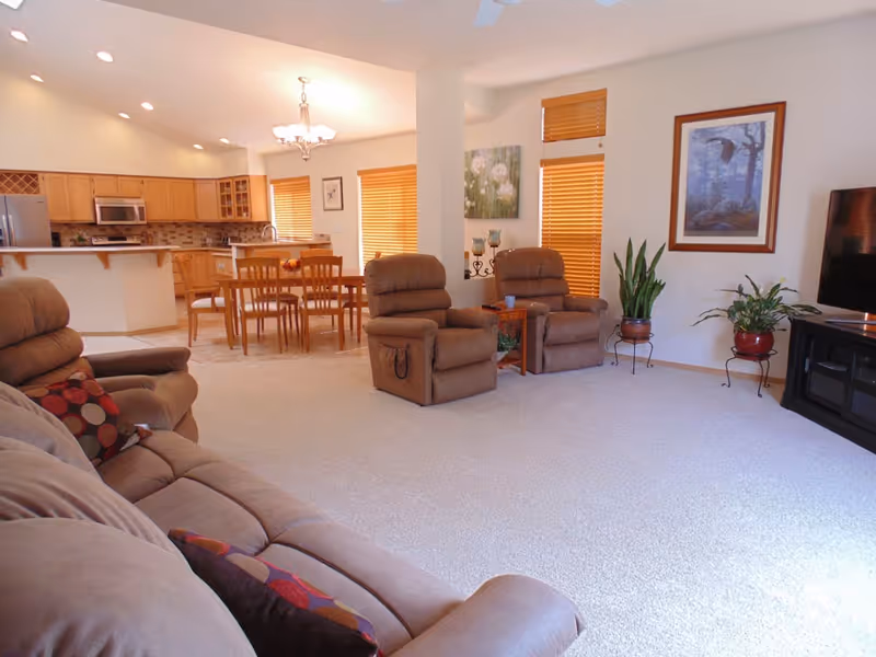 A spacious living room and dining area with beige carpet and walls. The living room has two brown recliner chairs, a brown sofa with patterned pillows, a flat-screen TV on a black stand, and several potted plants. The dining area features a wooden dining table with six chairs and is adjacent to a kitchen with wooden cabinets, a stainless steel refrigerator, microwave, and a tiled backsplash. The room is well-lit with natural light coming through windows with wooden blinds.