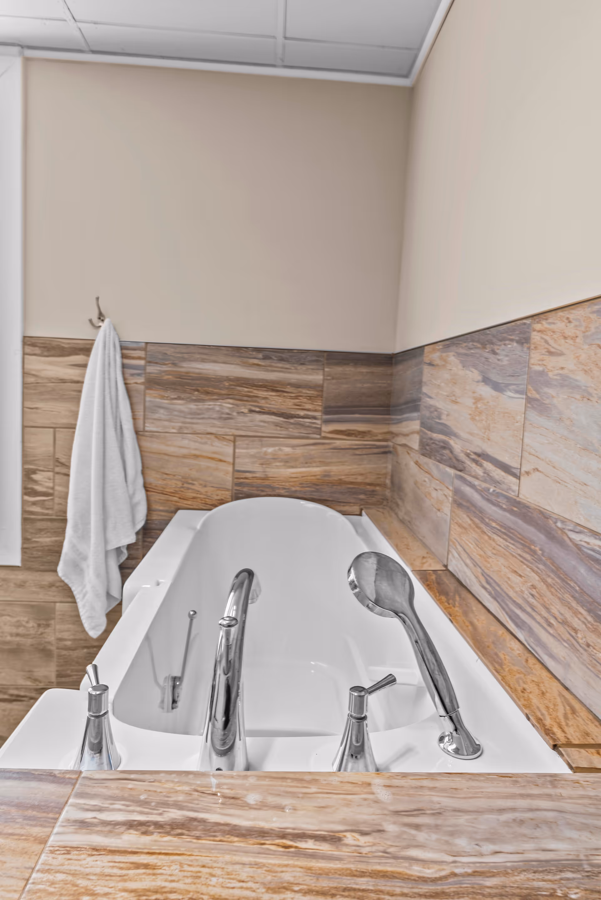 A white bathtub with chrome faucet and handheld showerhead, surrounded by beige and brown marble tiles on the walls and ledge. A white towel hangs on a hook on the wall to the left.