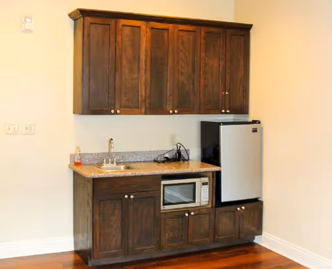 Small kitchenette with dark wood cabinets, a granite countertop, sink, microwave, and mini fridge against a light-colored wall.