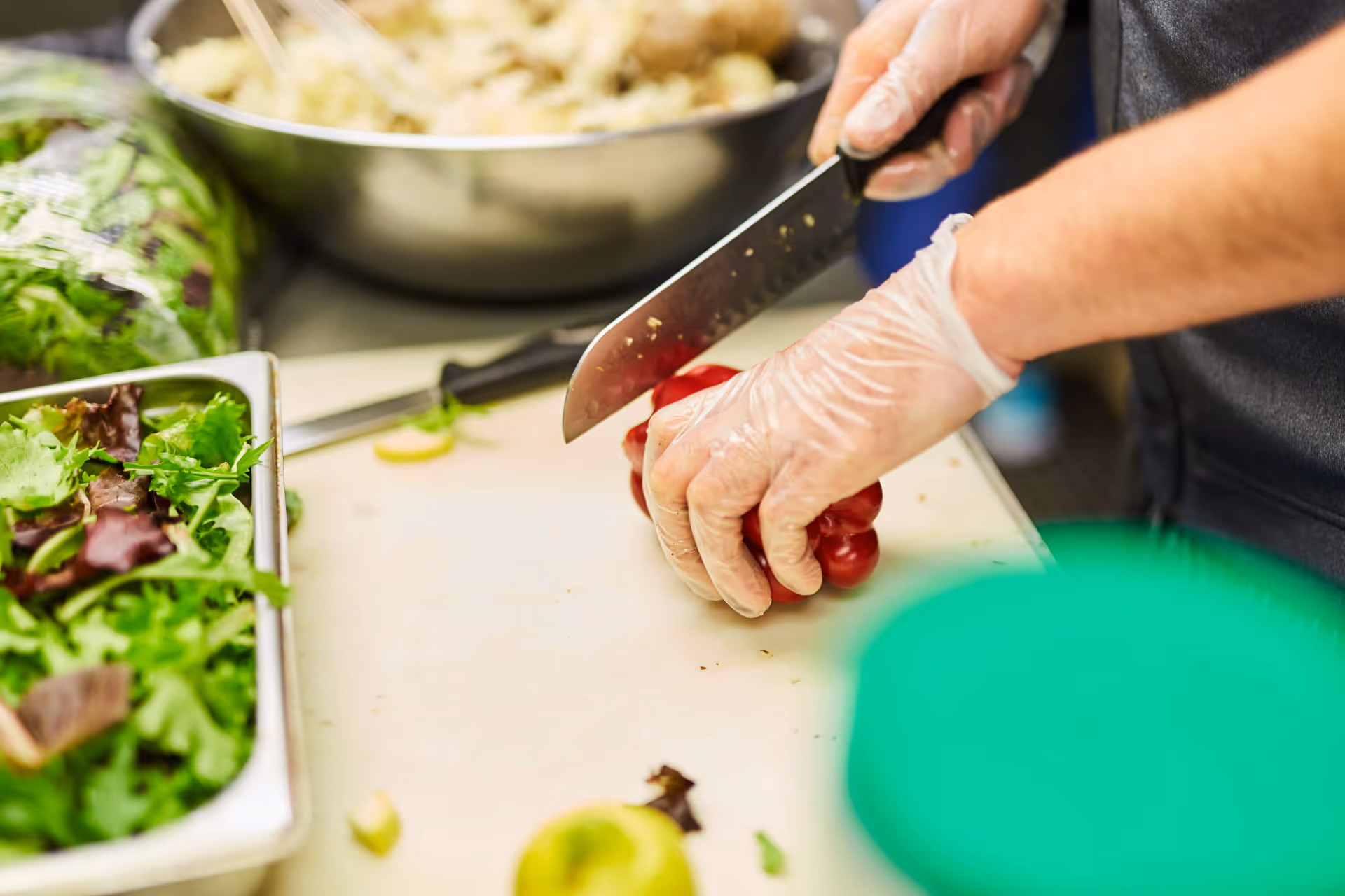 A person wearing disposable gloves is slicing a red bell pepper on a cutting board in a kitchen setting. There is a tray of mixed greens and a large metal bowl with food in the background.