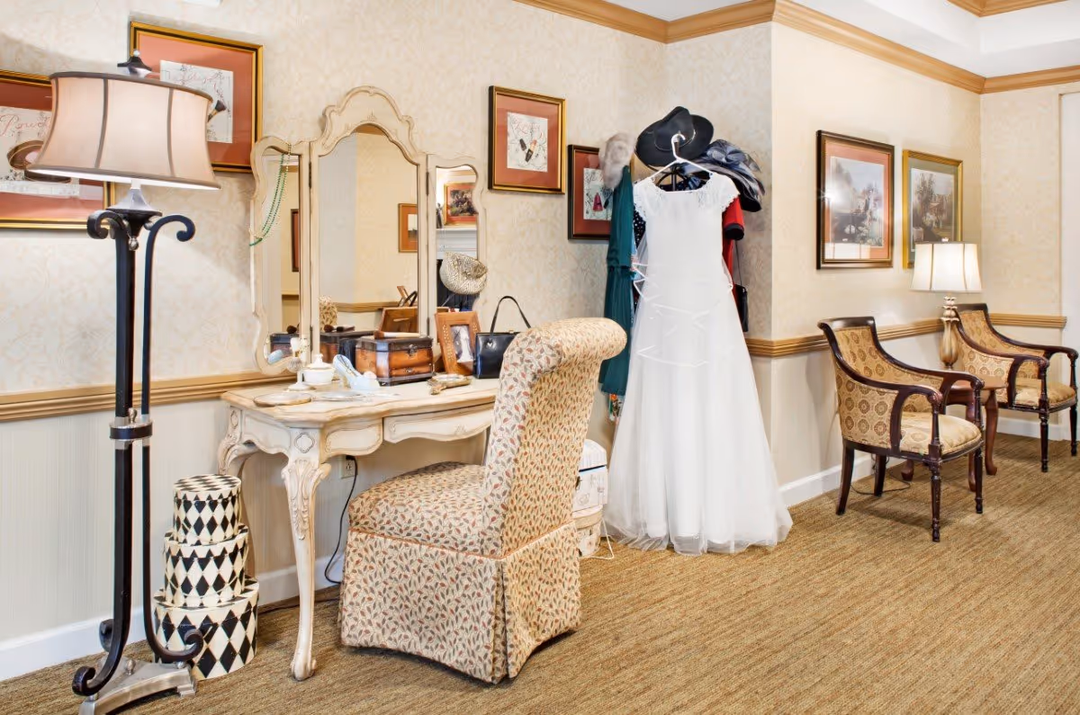 A cozy interior corner featuring a vintage-style vanity table with a tri-fold mirror, a cushioned chair with patterned upholstery, a floor lamp, and a coat rack holding a white dress and several hats. The walls are decorated with framed artwork, and two upholstered armchairs with wooden arms sit against the wall under a table lamp.