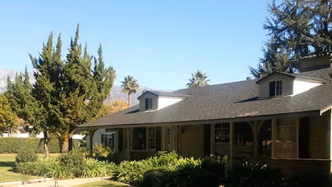 Exterior view of a single-story building with a dark shingled roof, multiple windows, and a covered porch area. The building is surrounded by green shrubs, trees, and a well-maintained lawn under a clear blue sky.