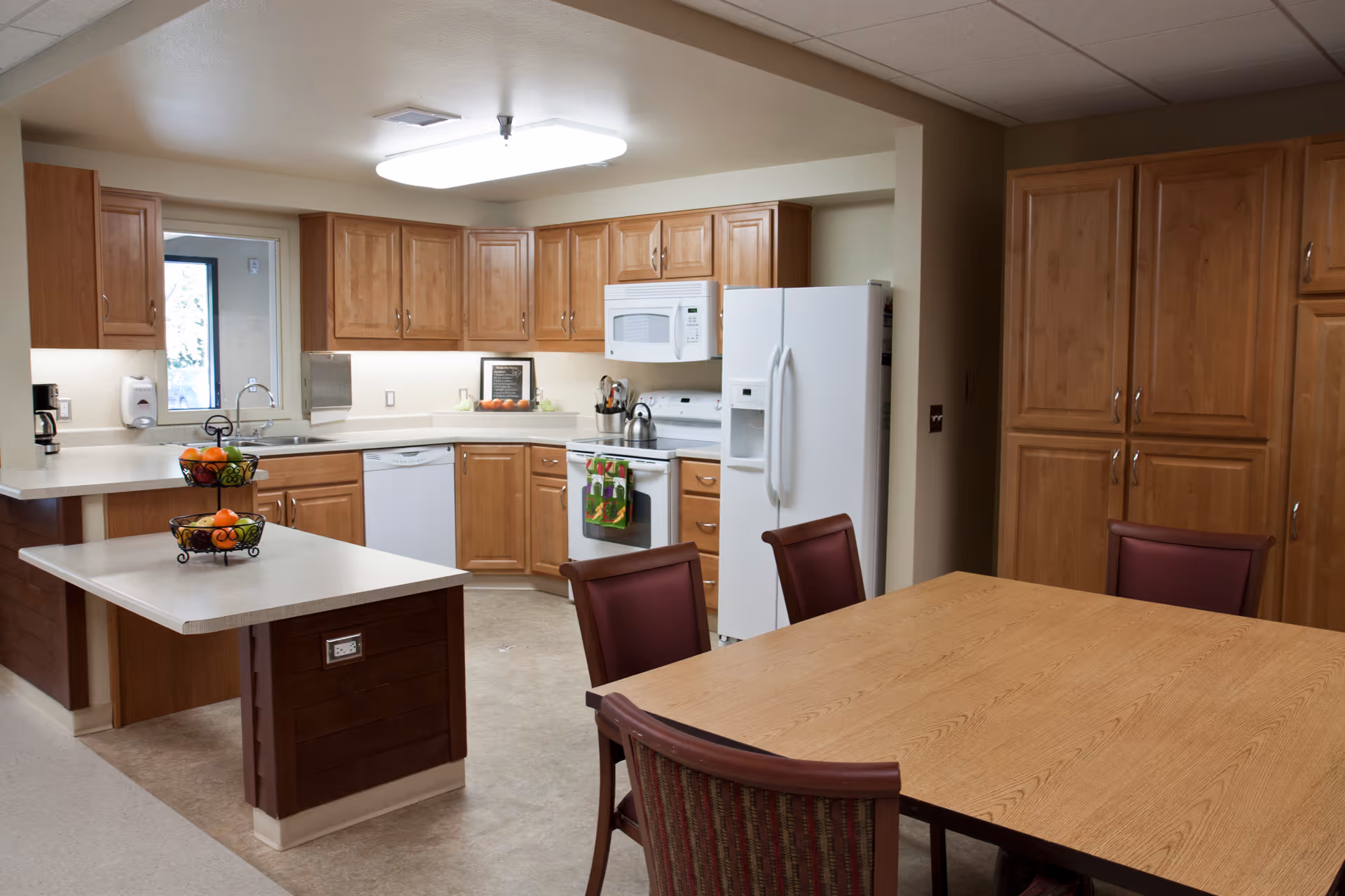 A clean and well-lit kitchen area with wooden cabinets, a white refrigerator, microwave, stove, dishwasher, and a sink. There is a small island with a two-tiered fruit basket and a dining table with four chairs in the foreground.