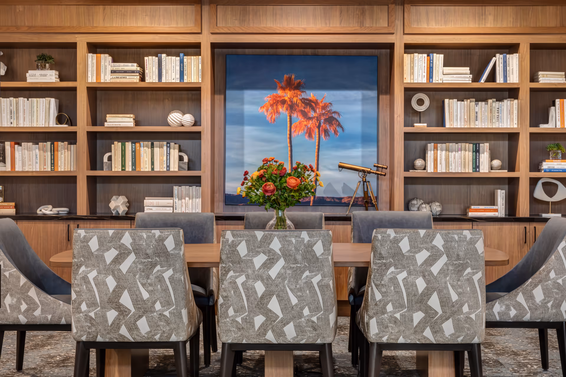 A modern dining room with a wooden table surrounded by eight patterned upholstered chairs. Behind the table is a large built-in wooden bookshelf filled with books and decorative objects. A colorful painting of palm trees and a telescope are displayed on the back wall. A vase with a bouquet of flowers is placed in the center of the table.