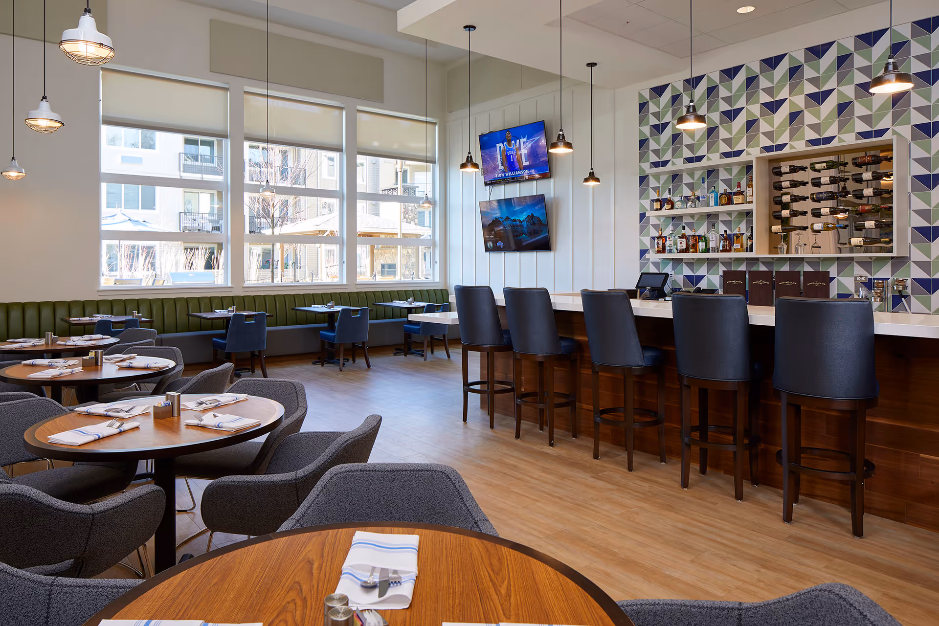 A modern dining area with round wooden tables set with napkins and silverware, gray upholstered chairs, a green cushioned bench along the windowed wall, and a bar with high black chairs in front of a geometric patterned wall with shelves holding bottles and wine racks. Two flat-screen TVs are mounted on the wall above the bar.