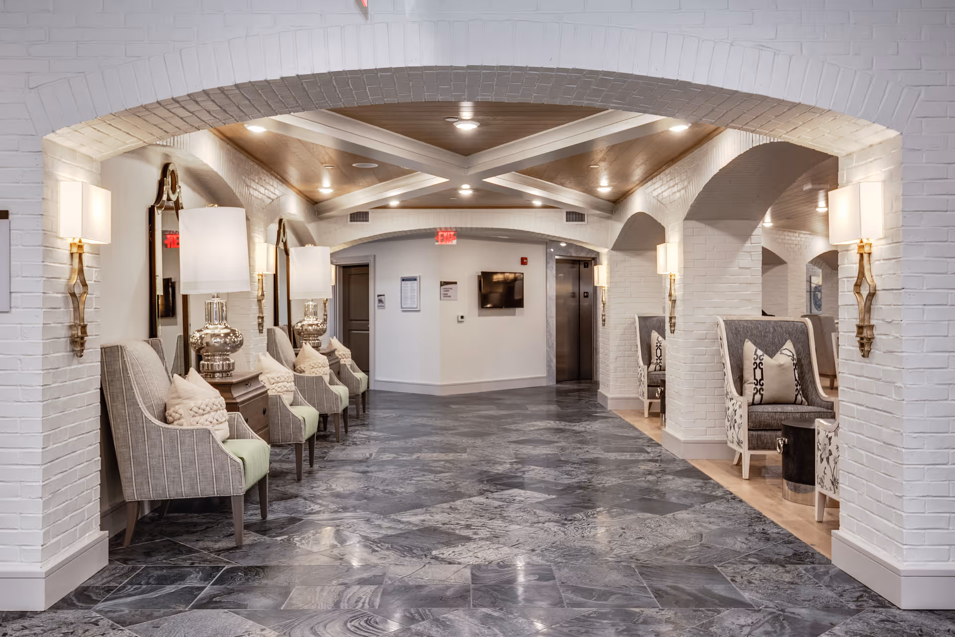 A spacious, well-lit hallway in a senior living facility with white brick walls and arched ceilings. The floor is covered with gray marble tiles. On the left side, there are several cushioned armchairs with pillows, placed in front of large mirrors and decorative lamps on wooden tables. On the right side, there are more cushioned chairs with patterned upholstery and small tables. At the end of the hallway, there is an elevator and a wall-mounted TV.