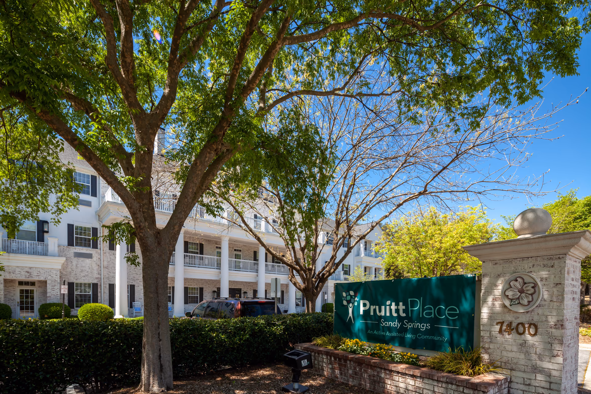Exterior view of PruittPlace - Sandy Springs, an assisted living community, showing a large white building with multiple windows and balconies. In the foreground, there is a green sign with the facility's name and address number 7400 on a brick pillar. Trees and bushes surround the area under a clear blue sky.